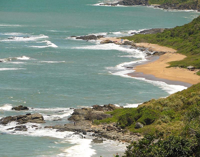 Vista aérea de uma praia de areia que se estende ao longo de uma costa verdejante, com ondas turquesas do oceano quebrando nas rochas.