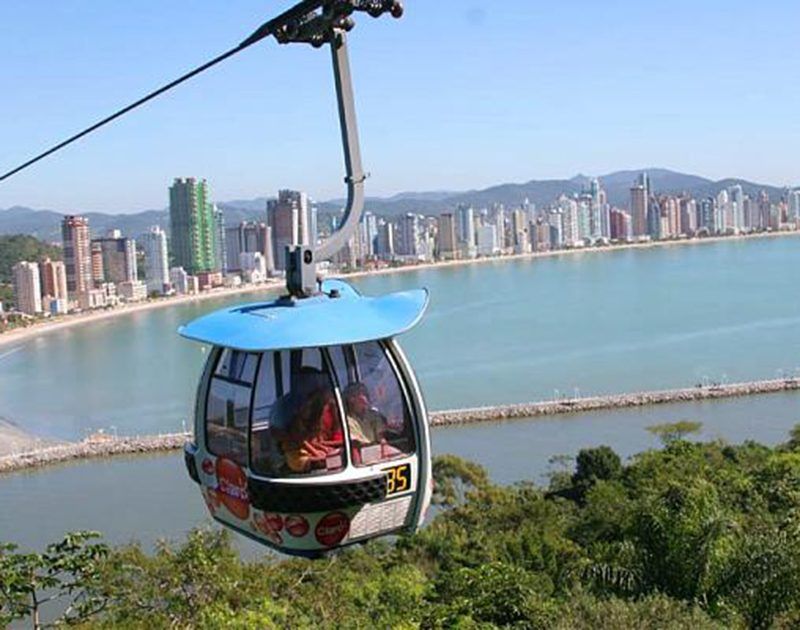 Um teleférico com dois passageiros suspenso acima de uma praia urbana com arranha-céus e água azul cristalina.