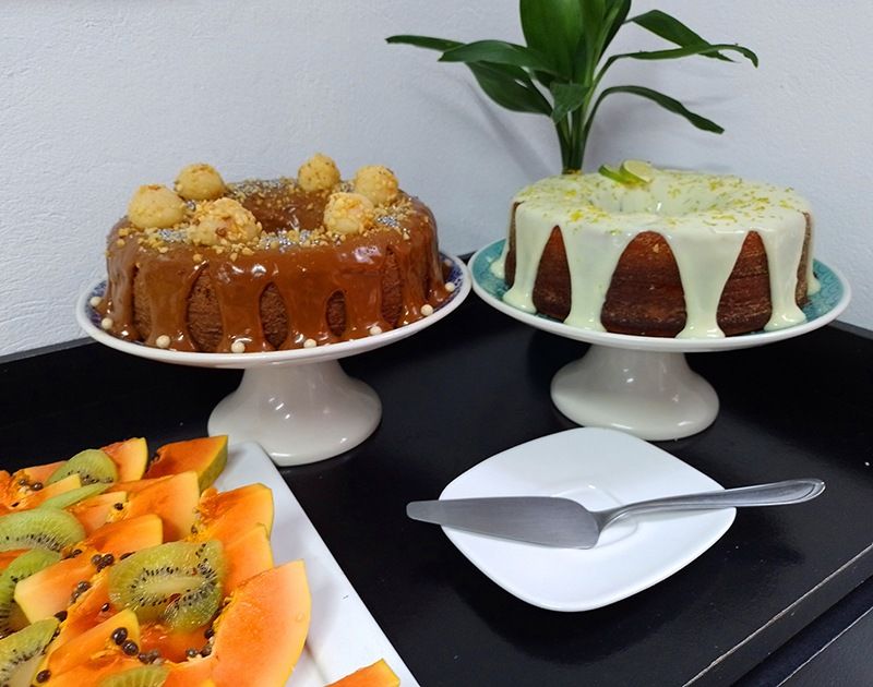 Dois bolos Bundt em suportes, um com cobertura de caramelo e outro com glacê branco, ao lado de um prato de frutas fatiadas.