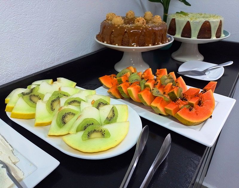 Uma mesa de bufê com travessas de melão, kiwi e mamão fatiados, ao lado de dois bolos em pedestais.