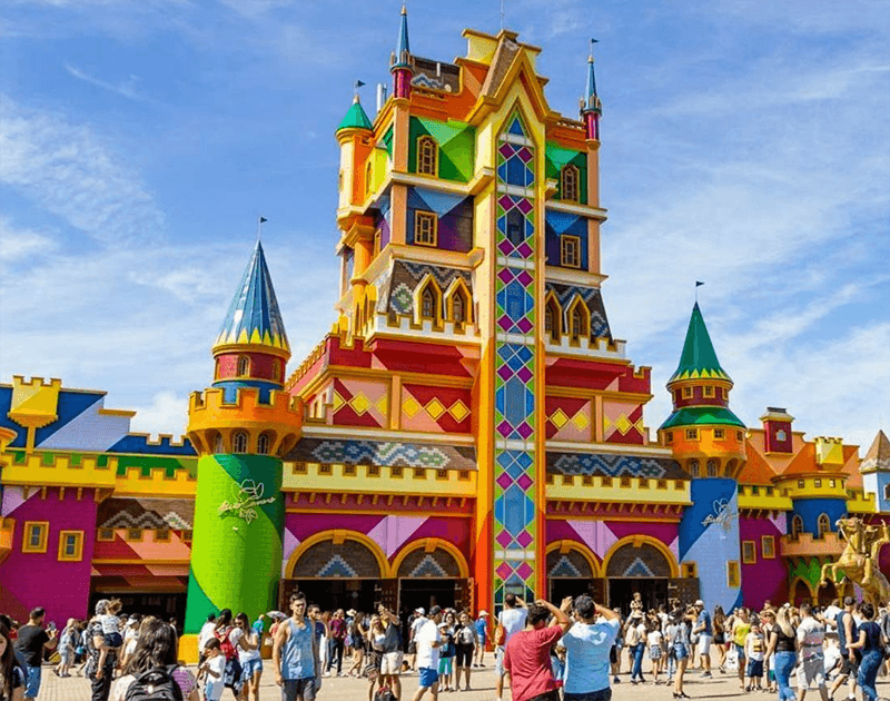 Um castelo colorido e extravagante no parque temático Beto Carrero World, com multidões de pessoas caminhando em frente em um dia ensolarado.