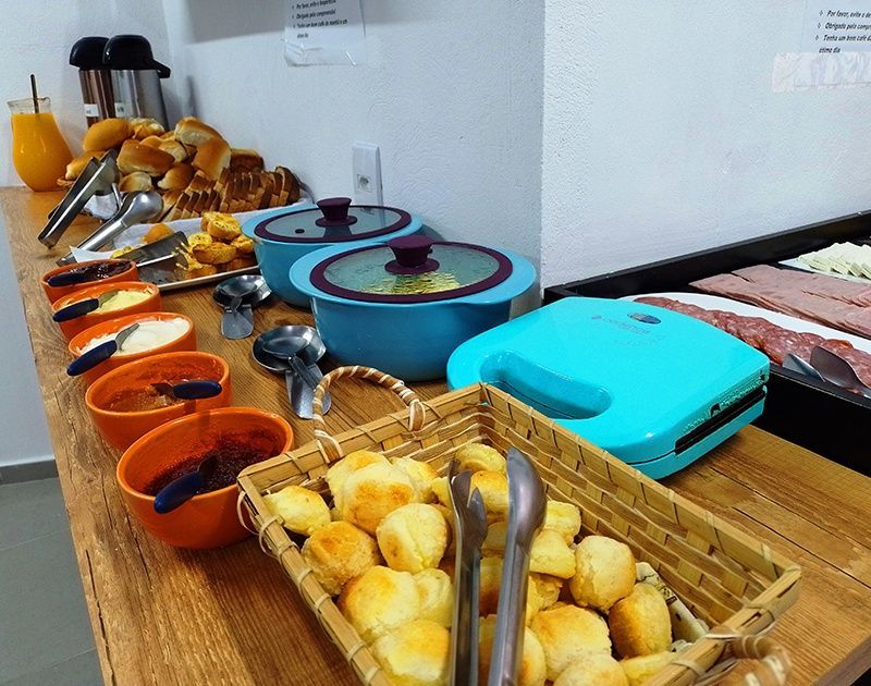 Um balcão de buffet de café da manhã com tigelas de ingredientes, uma cesta de pãezinhos de queijo, uma prensa de panini e pães variados.