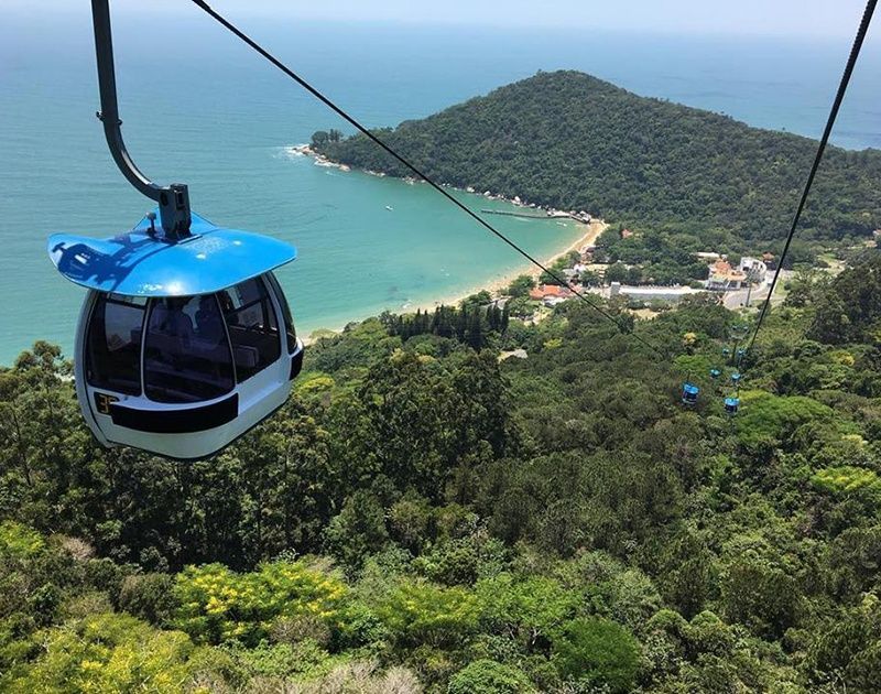 Um teleférico azul desliza sobre uma exuberante floresta verde, com uma praia de areia e o oceano azul visíveis ao fundo.