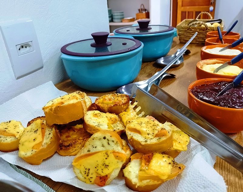 Uma pilha de pão torrado coberto com queijo derretido ao lado de várias pequenas tigelas coloridas com pastas, sobre uma mesa de madeira.