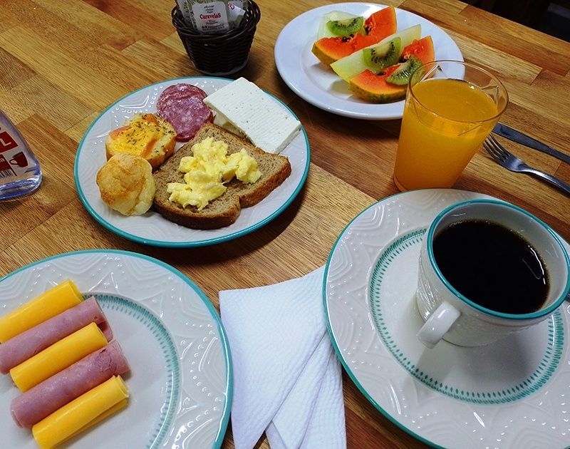 Uma mesa posta com itens de café da manhã: pratos de frutas, ovos mexidos na torrada, queijo, salame, presunto, suco e café preto.