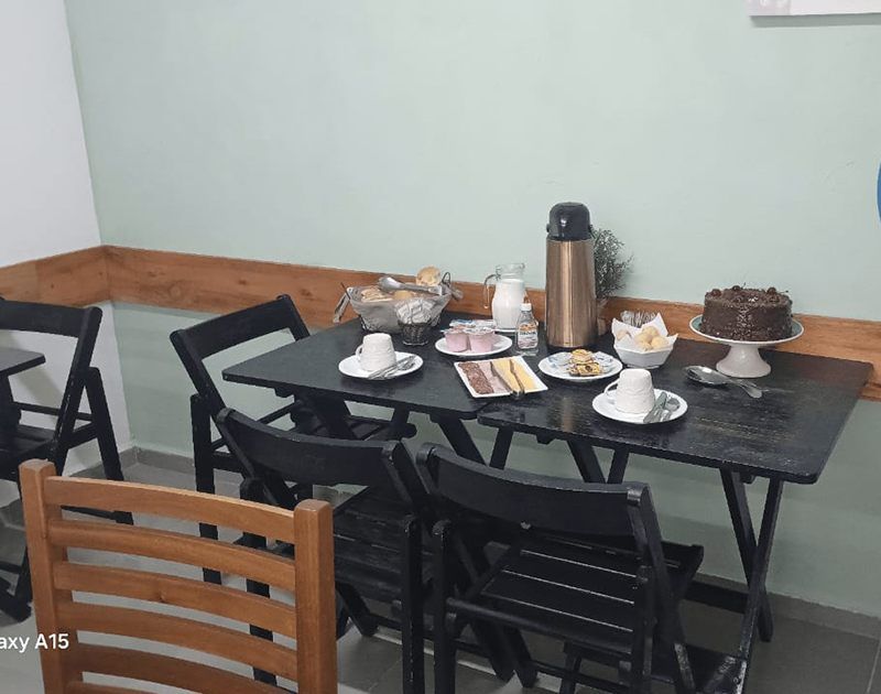 Uma mesa posta para café com um bolo, uma garrafa térmica, pão, pratos e xícaras em uma sala com paredes verde-claras.