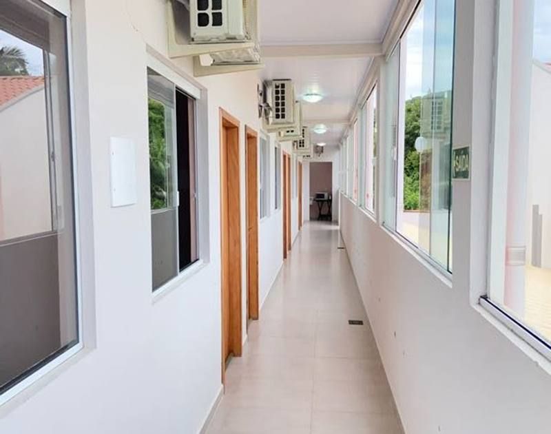Um corredor longo e bem iluminado, com paredes brancas, portas de madeira, janelas de vidro e aparelhos de ar condicionado instalados.