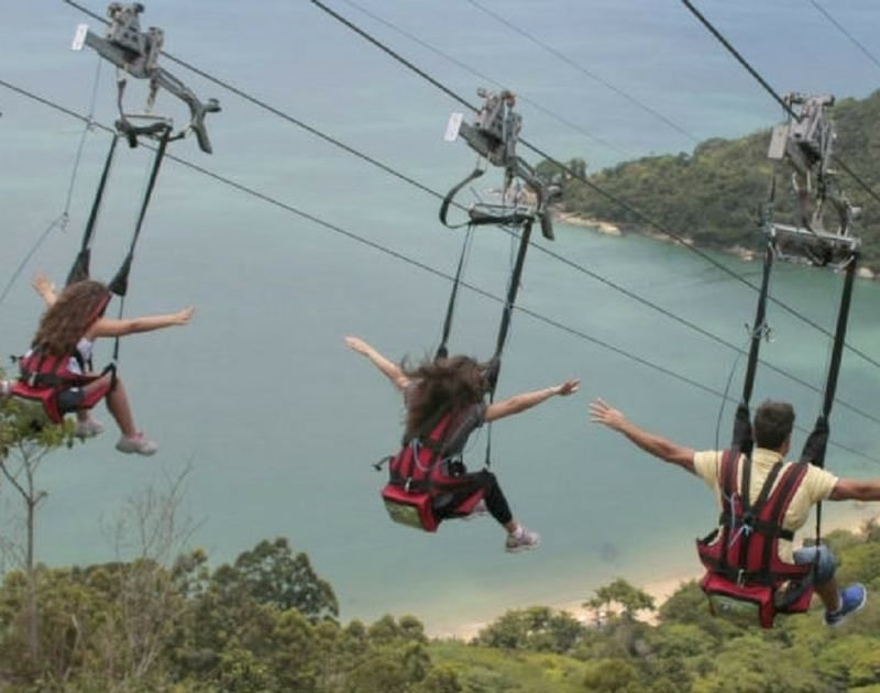 Três pessoas suspensas por arneses deslizam em tirolesas paralelas sobre uma costa panorâmica com árvores e praias de areia.