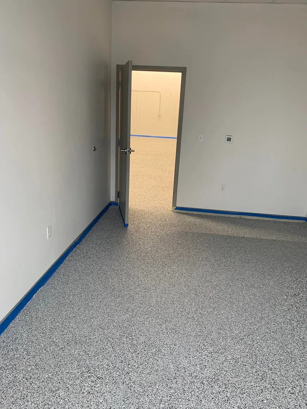Empty room with speckled floor, white walls, blue baseboards, and an open door to a sunlit area.