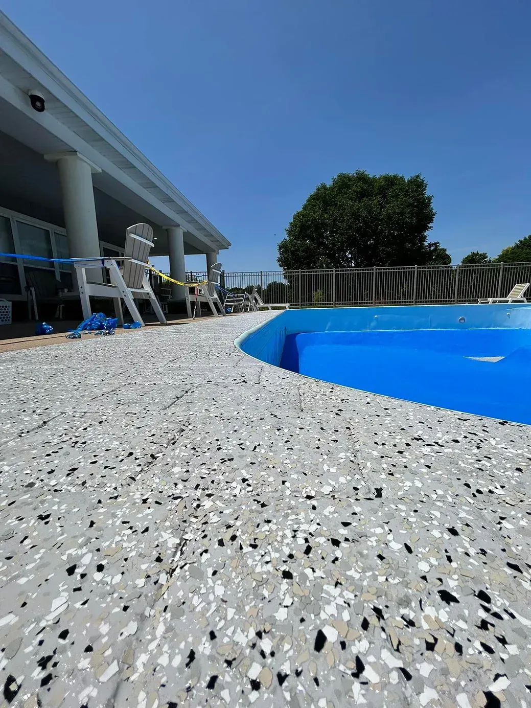 Low-angle view of a blue swimming pool beside a house with chairs and a tree on a sunny day