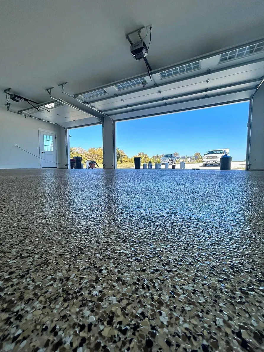 Empty garage interior with open door, rough speckled floor, and bright outdoor view beyond.