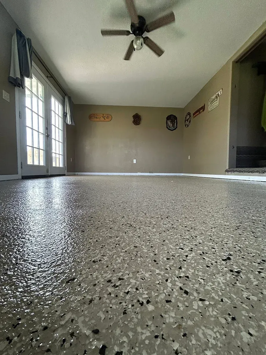 Empty living room with speckled floor, ceiling fan, windows, and wall decor.