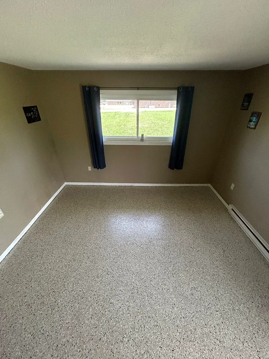 Empty beige room with speckled floor, window with dark curtains, and wall outlets on both sides