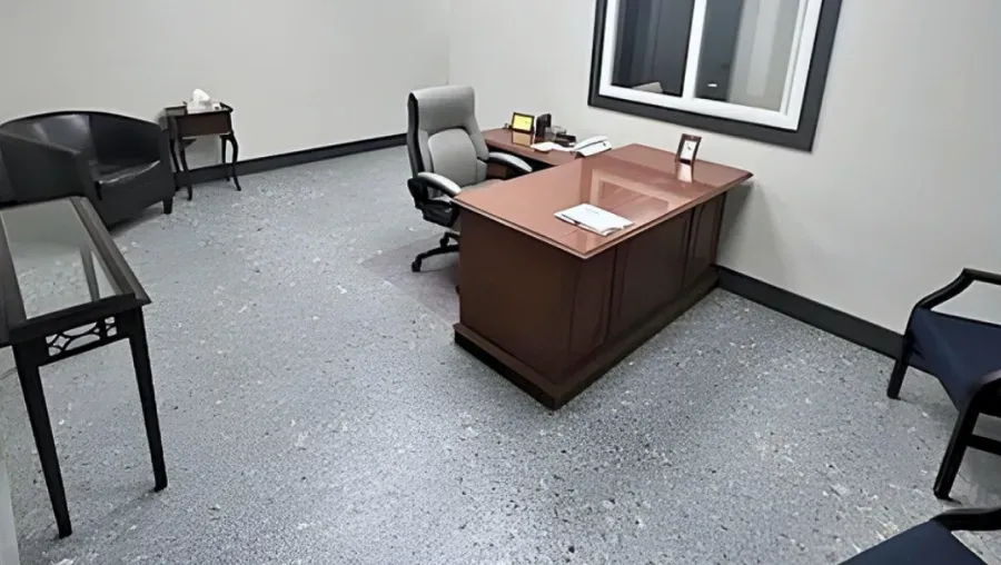 Small office with wooden desk, gray chair, black side table, and blue chairs on a speckled floor.