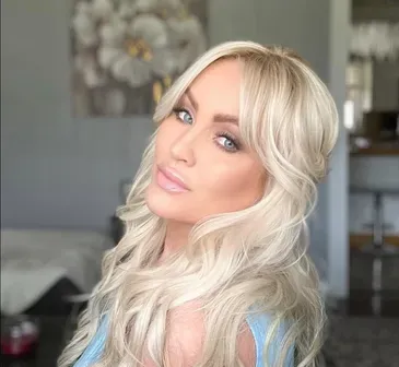 Blonde woman with long wavy hair, looking at the camera. She's in a room, with artwork on the wall.