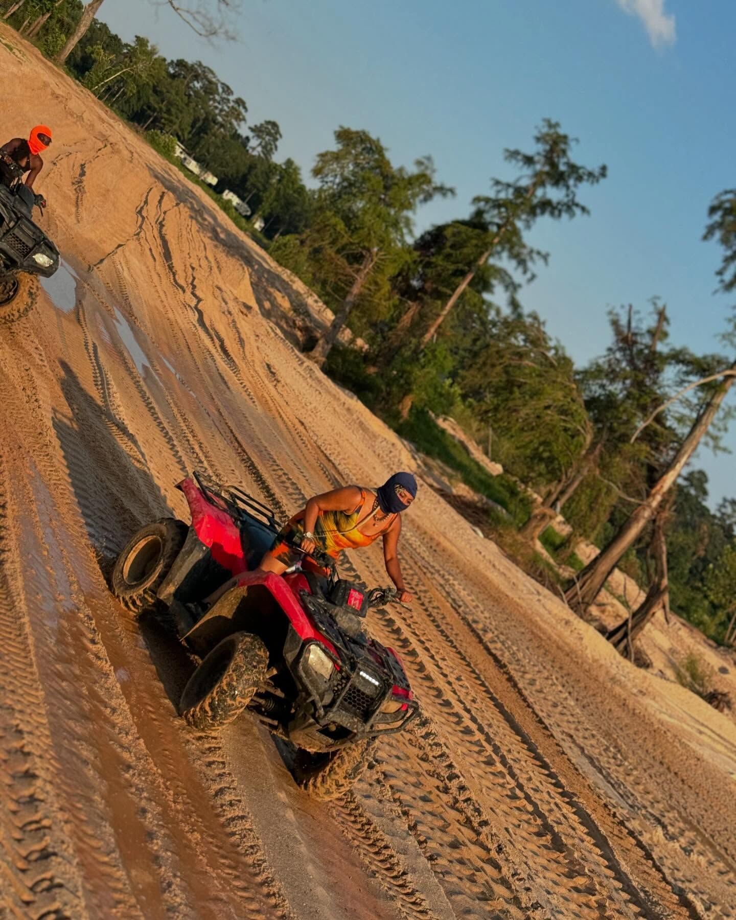 A person in a dark helmet and colorful shirt rides a red ATV through a muddy, tire-tracked, sunlit field with trees.