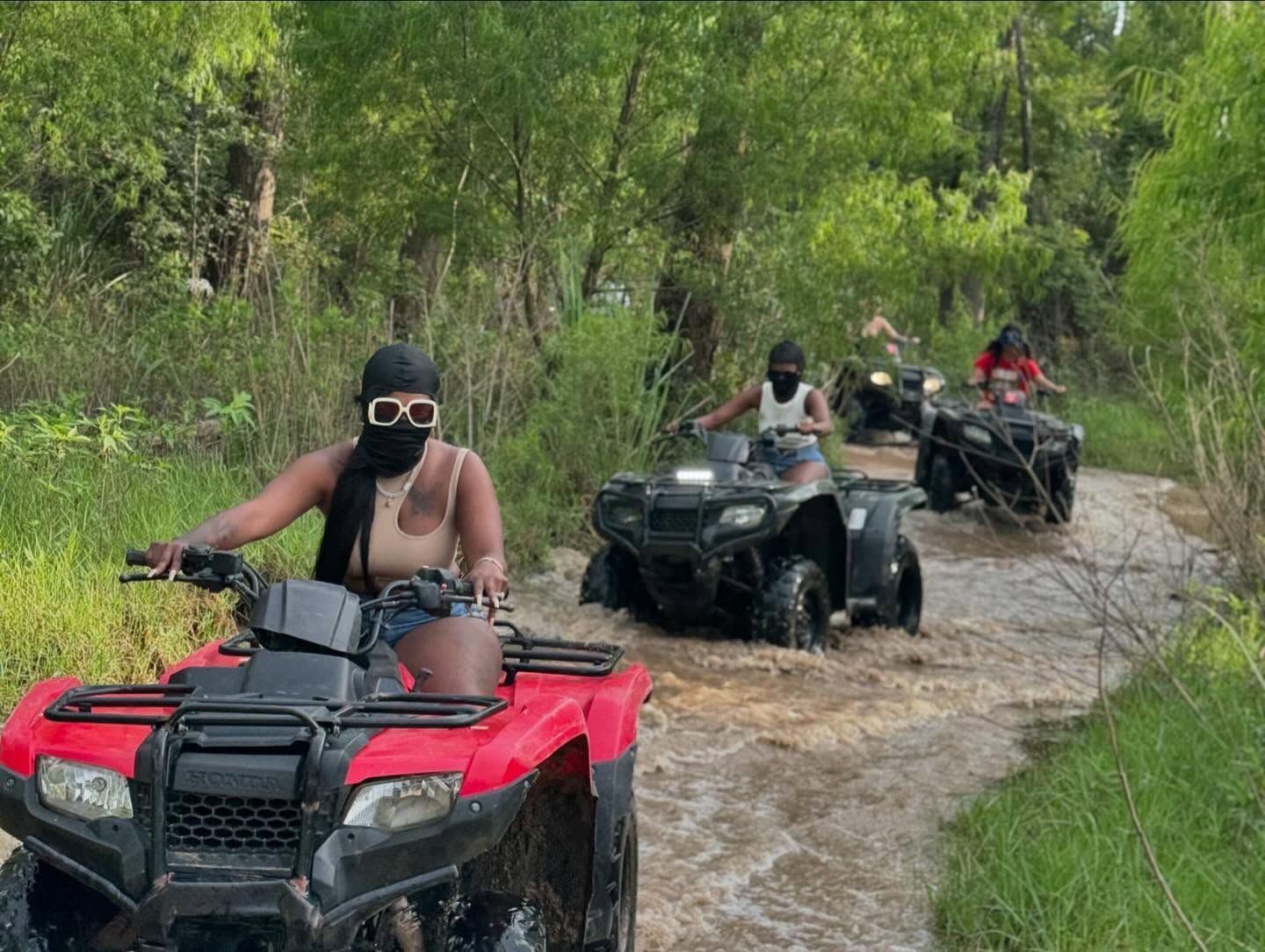 People riding four-wheelers through a muddy, forested trail, with the foreground rider wearing sunglasses and a mask.