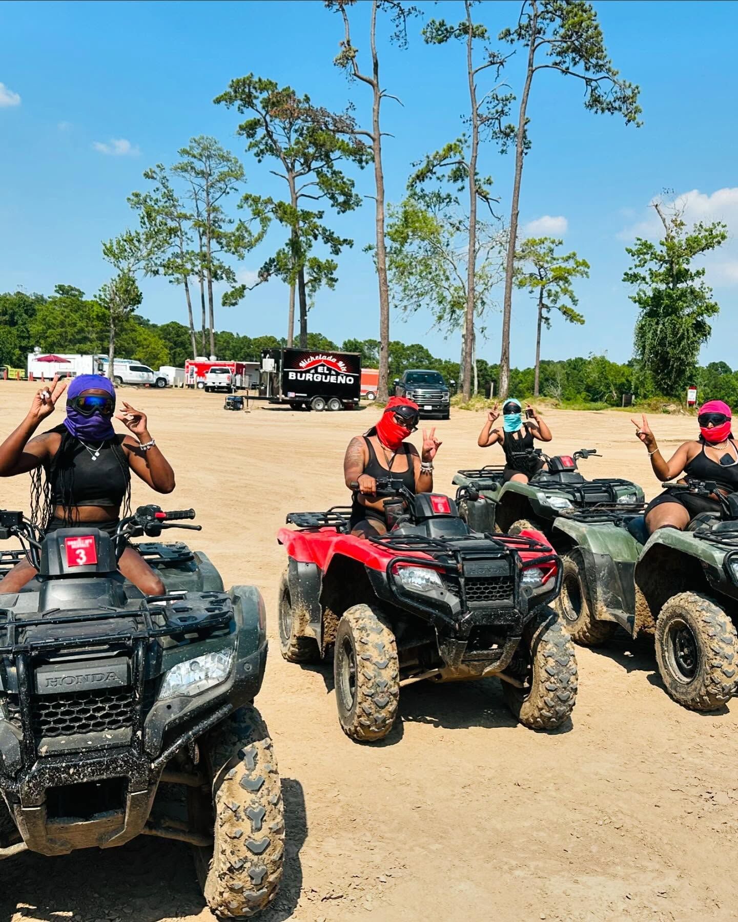 Four people wearing face coverings on ATVs in a sunny, sandy outdoor area with a truck and trailer in the background.