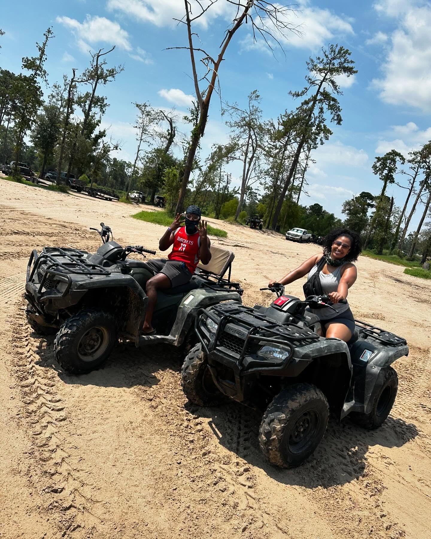 Two people sit on dark-colored ATVs in a sandy, outdoor trail setting with trees under a blue, partly cloudy sky.