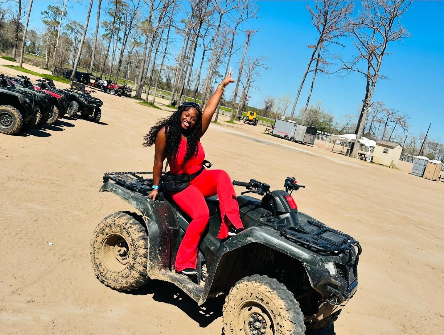 A person in a red outfit posing with one arm raised while sitting on a black ATV at an outdoor dirt lot.