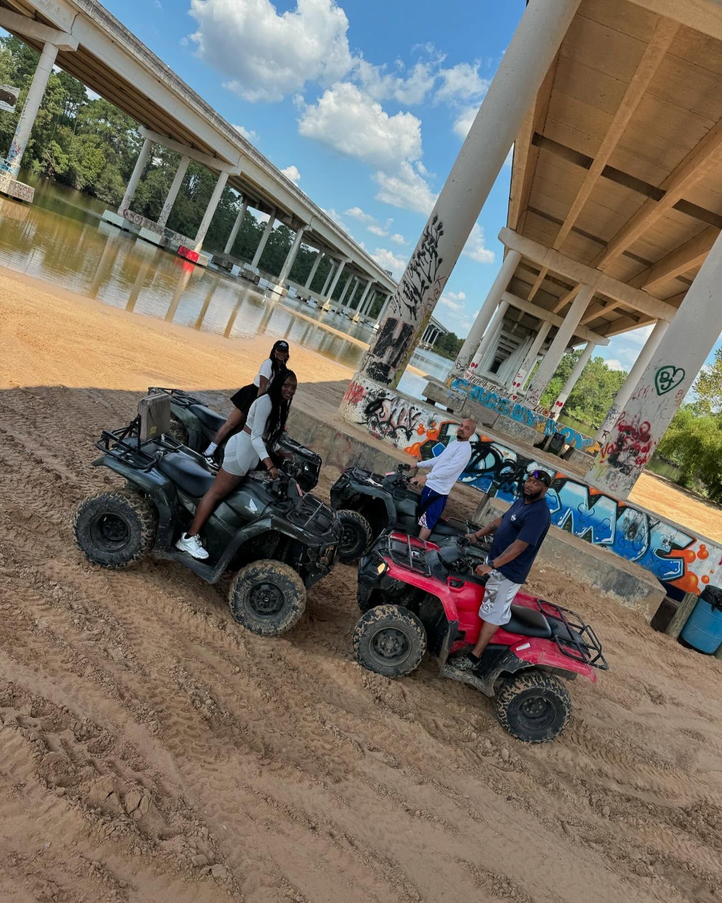 People sitting on ATVs on a sandy riverbank beneath a large concrete highway bridge on a sunny day.