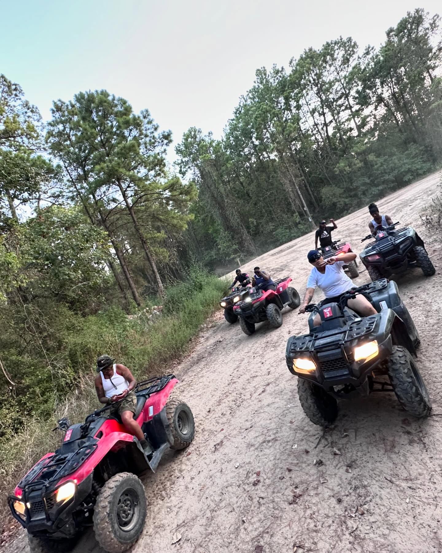 A group of people riding ATVs on a dirt trail surrounded by trees on a sunny day.