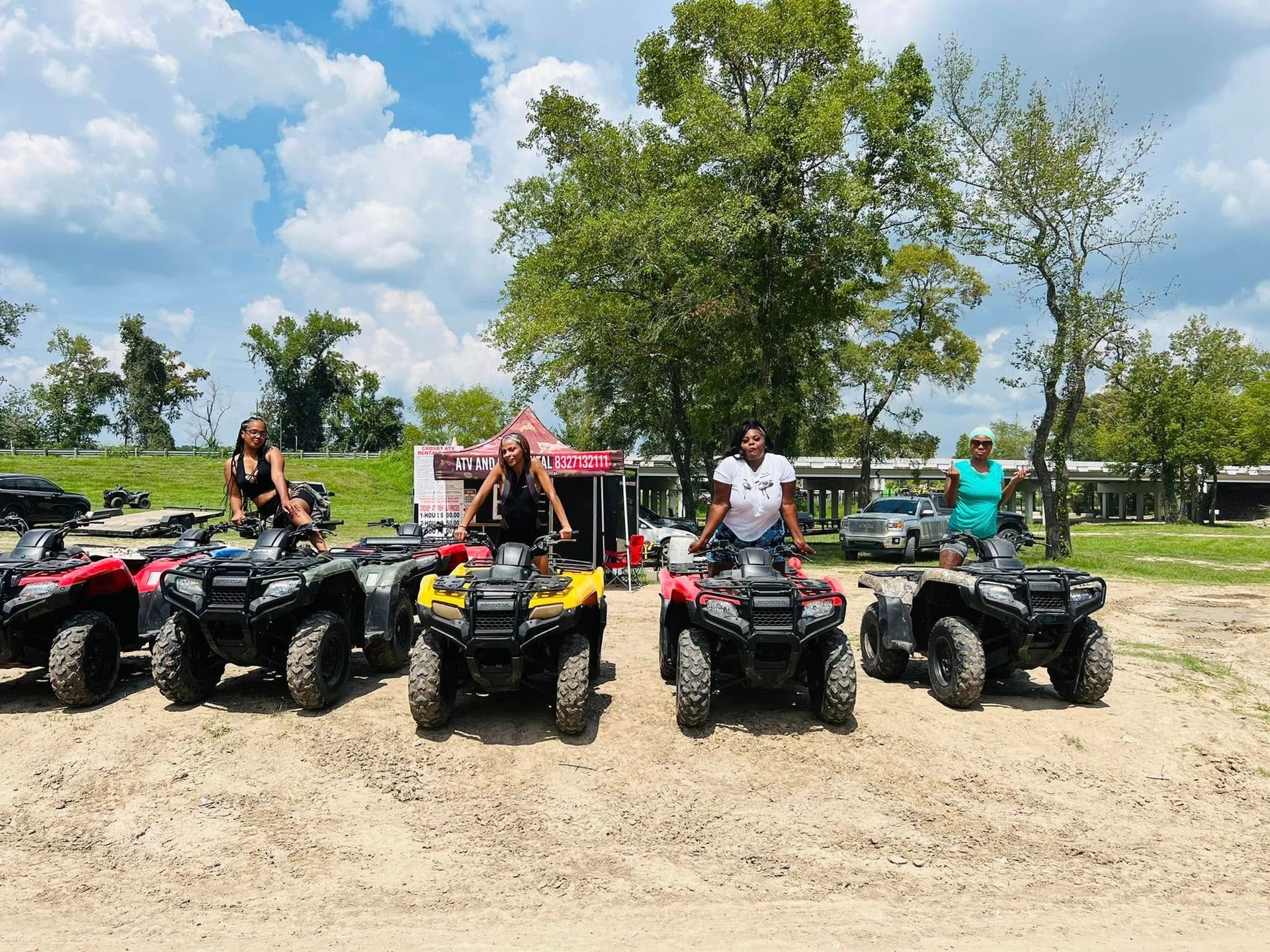 Four people posing on parked all-terrain vehicles in a grassy, outdoor area near a trailer under a blue, cloudy sky.