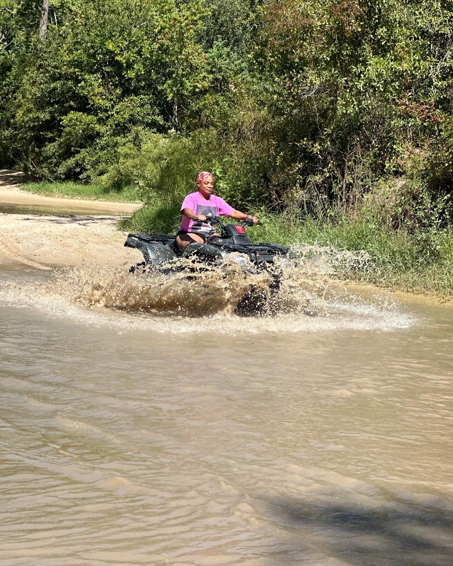 A person in a pink shirt rides an ATV through a shallow, muddy creek, creating a large splash.
