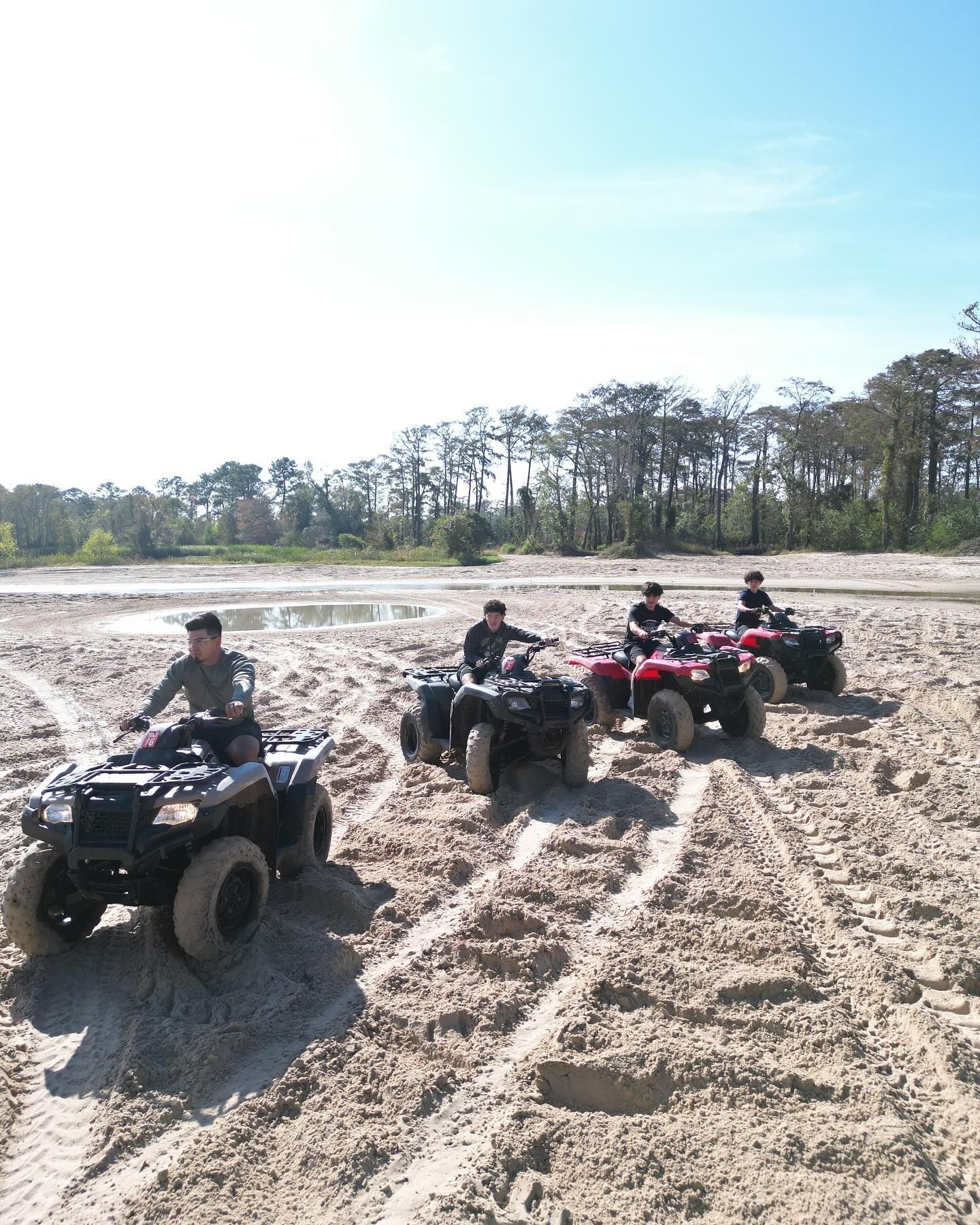 Four people ride ATVs across a sunny, sandy landscape with a line of trees in the background.