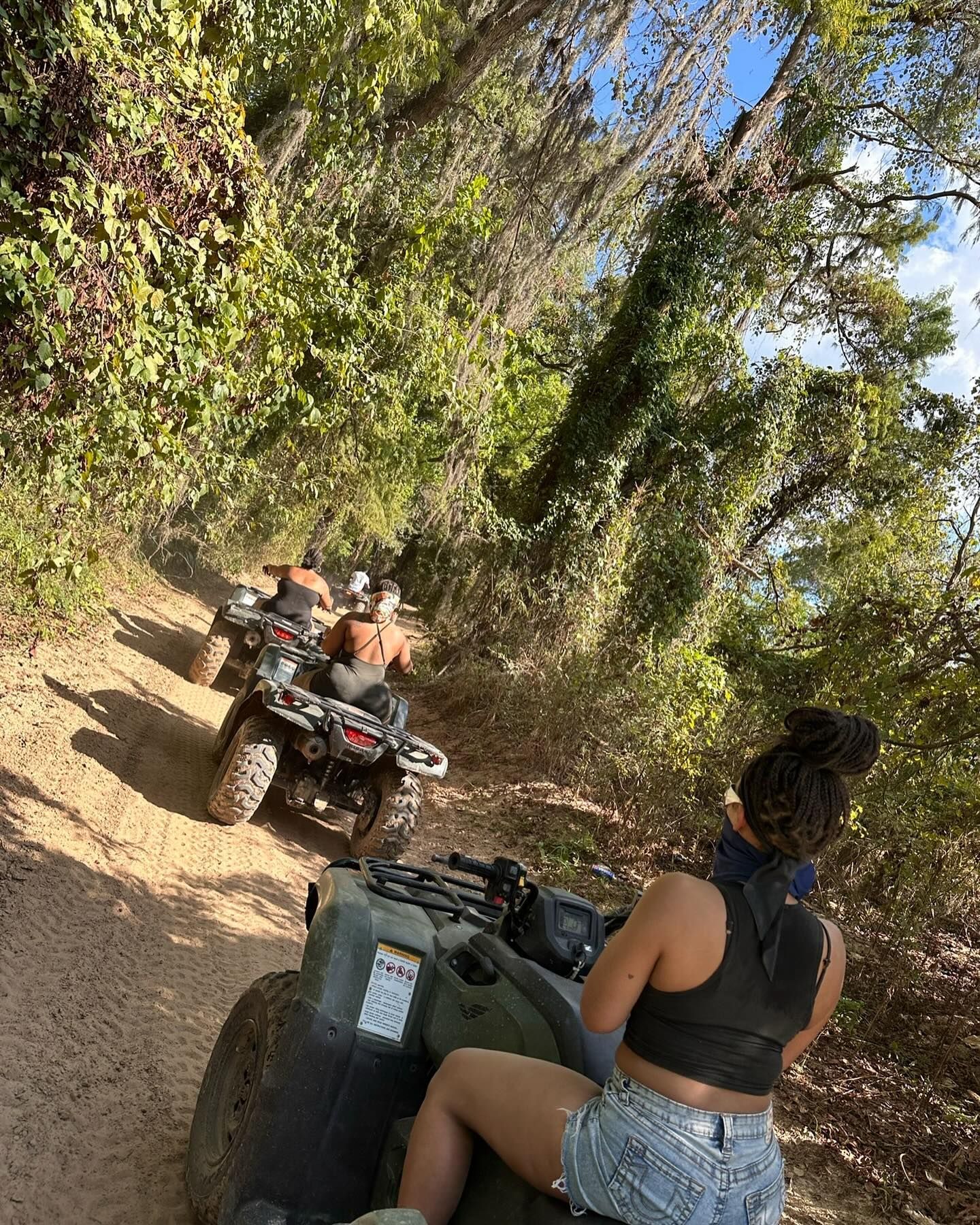 A group of people riding ATVs on a sunlit, tree-lined dirt trail in a wooded area.