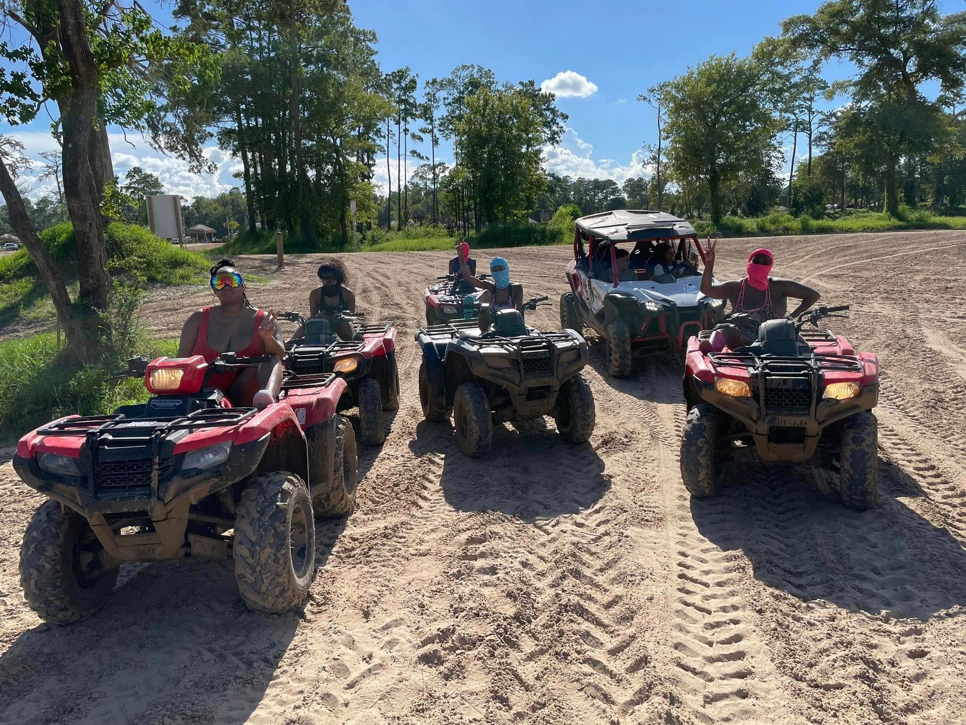 Four people riding ATVs and a UTV across a sandy, sunny off-road trail surrounded by trees.