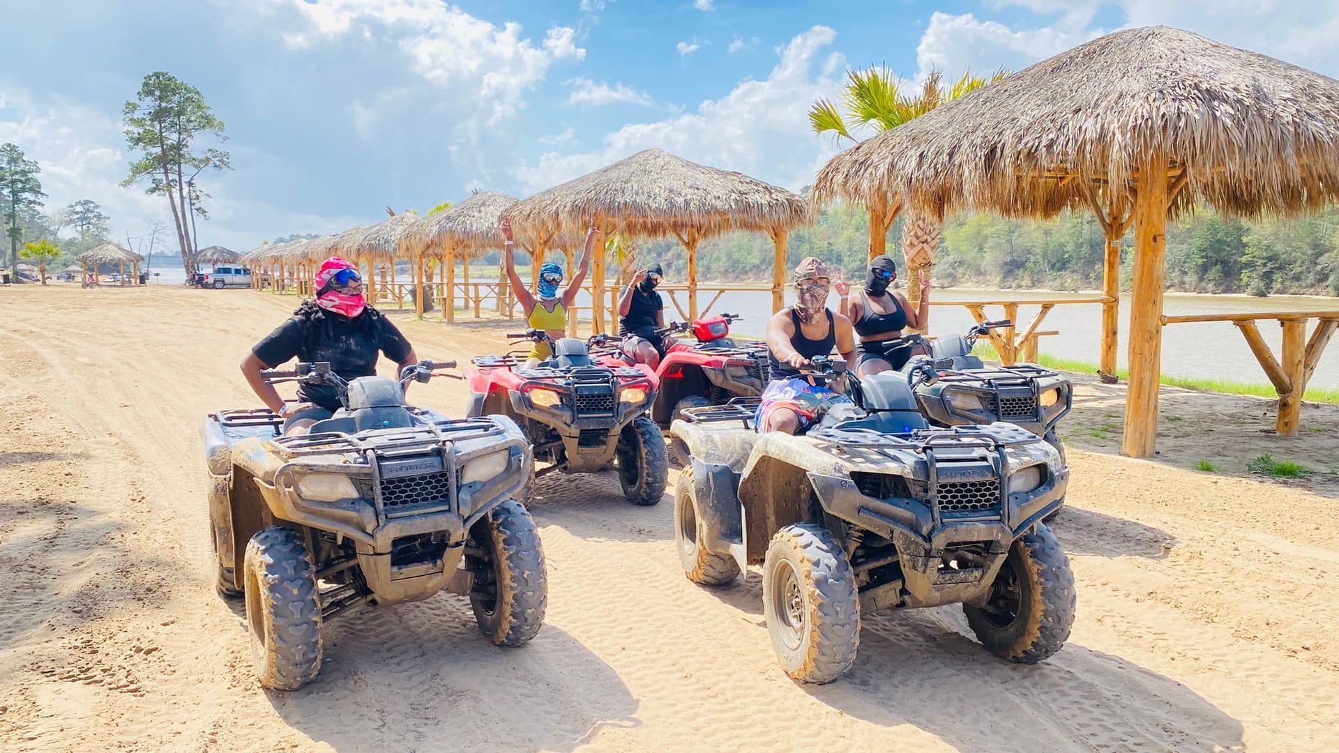 A group of people on ATVs posing in front of thatched-roof shelters on a sandy beach.