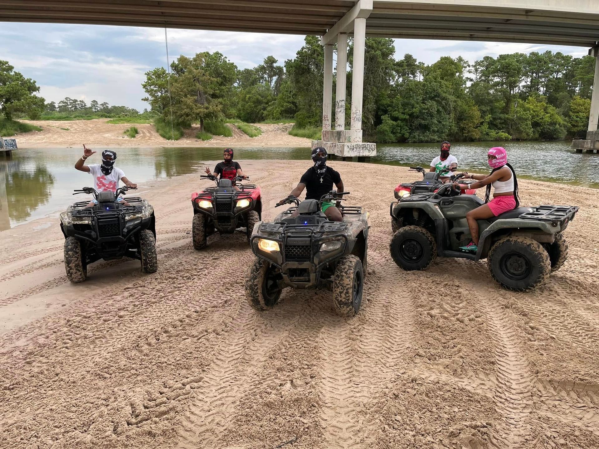 Five people ride ATVs on a sandy riverbank under a highway bridge, with one rider gesturing toward the camera.