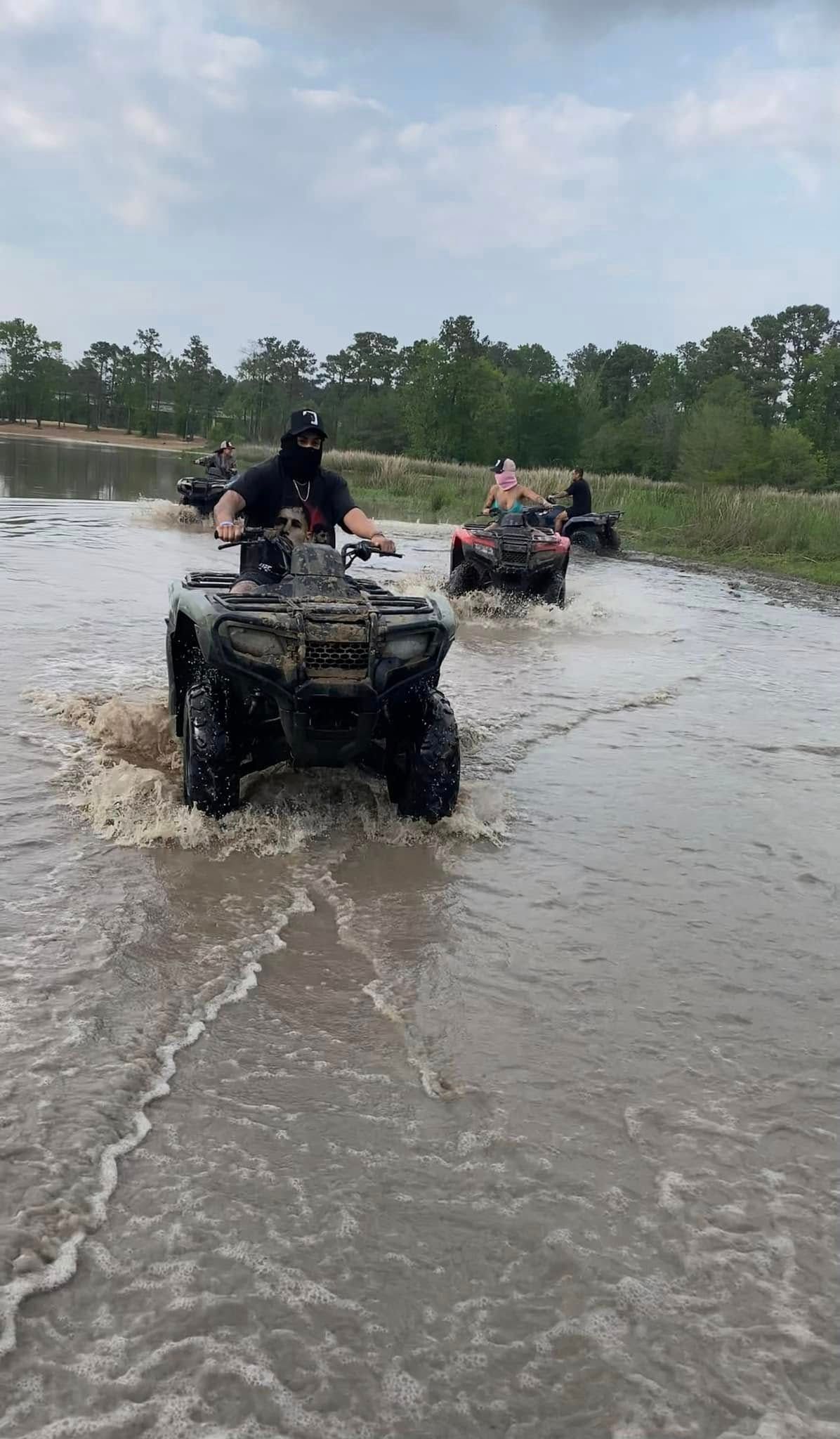 A group of people riding ATVs through a muddy body of water under a cloudy sky.