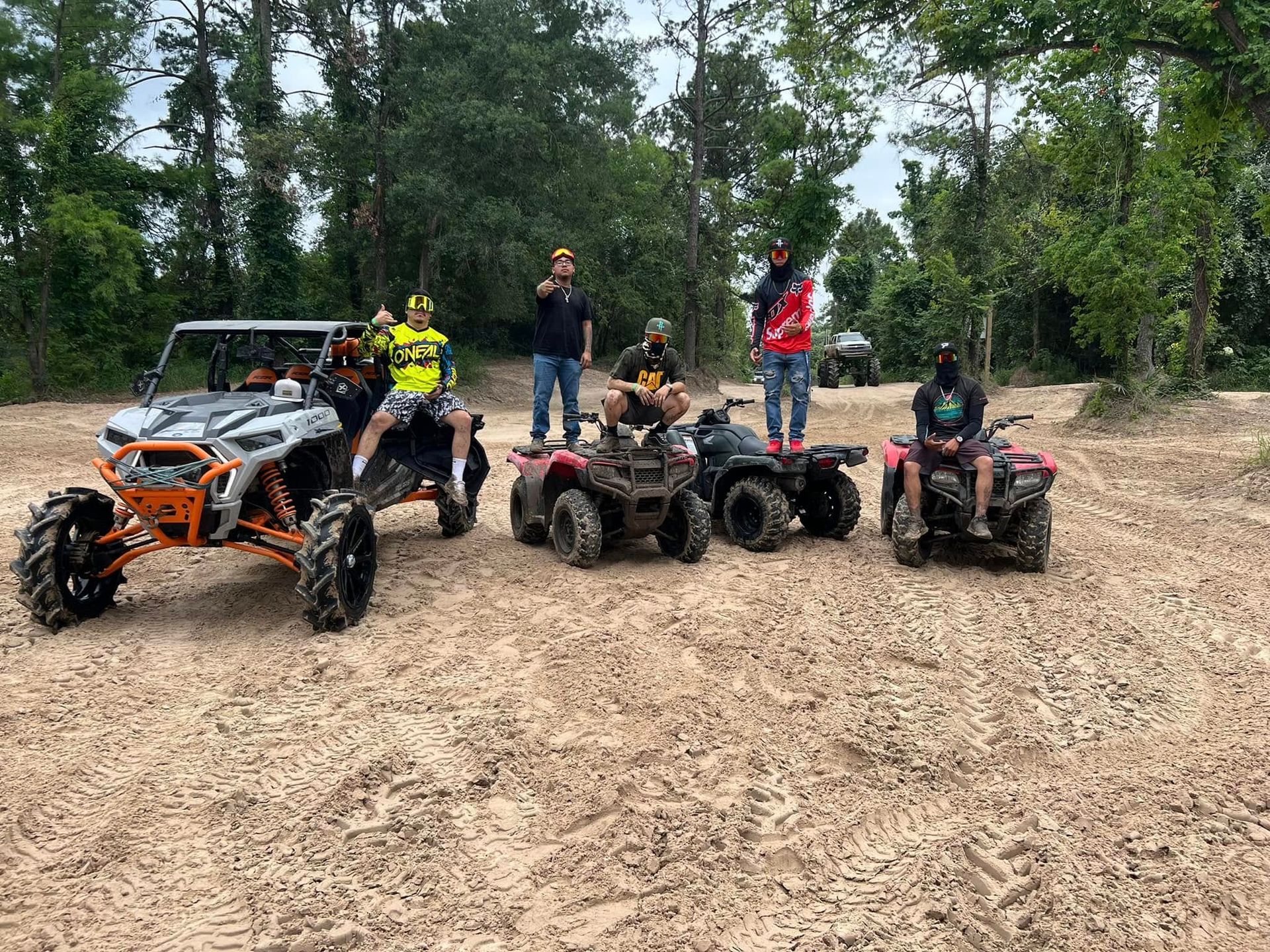 Group of people posing on ATVs and a UTV in a wooded, sandy off-road area.