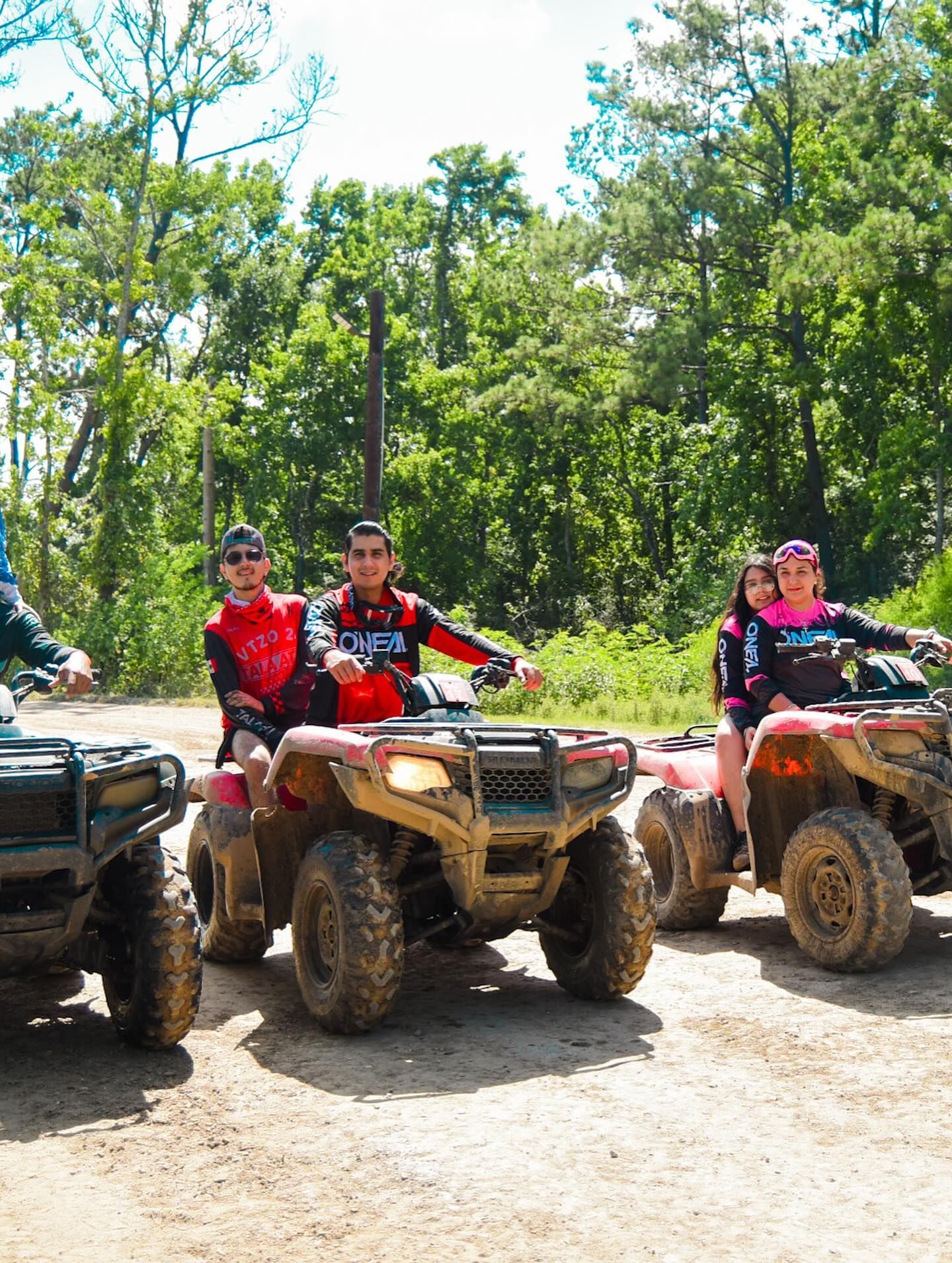 Three people riding ATVs on a dirt path in a wooded, sunny outdoor area.