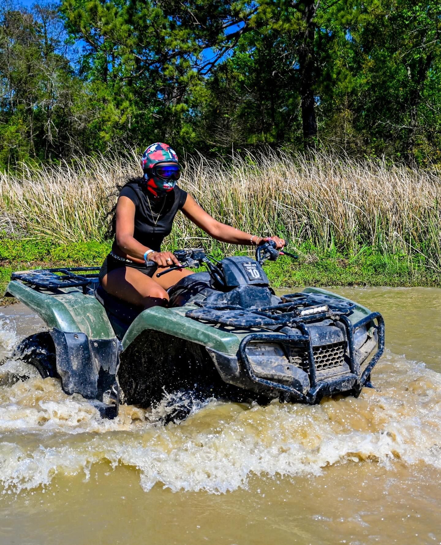 A person wearing a helmet and goggles rides a green ATV through shallow, muddy water in a grassy, wooded outdoor area.