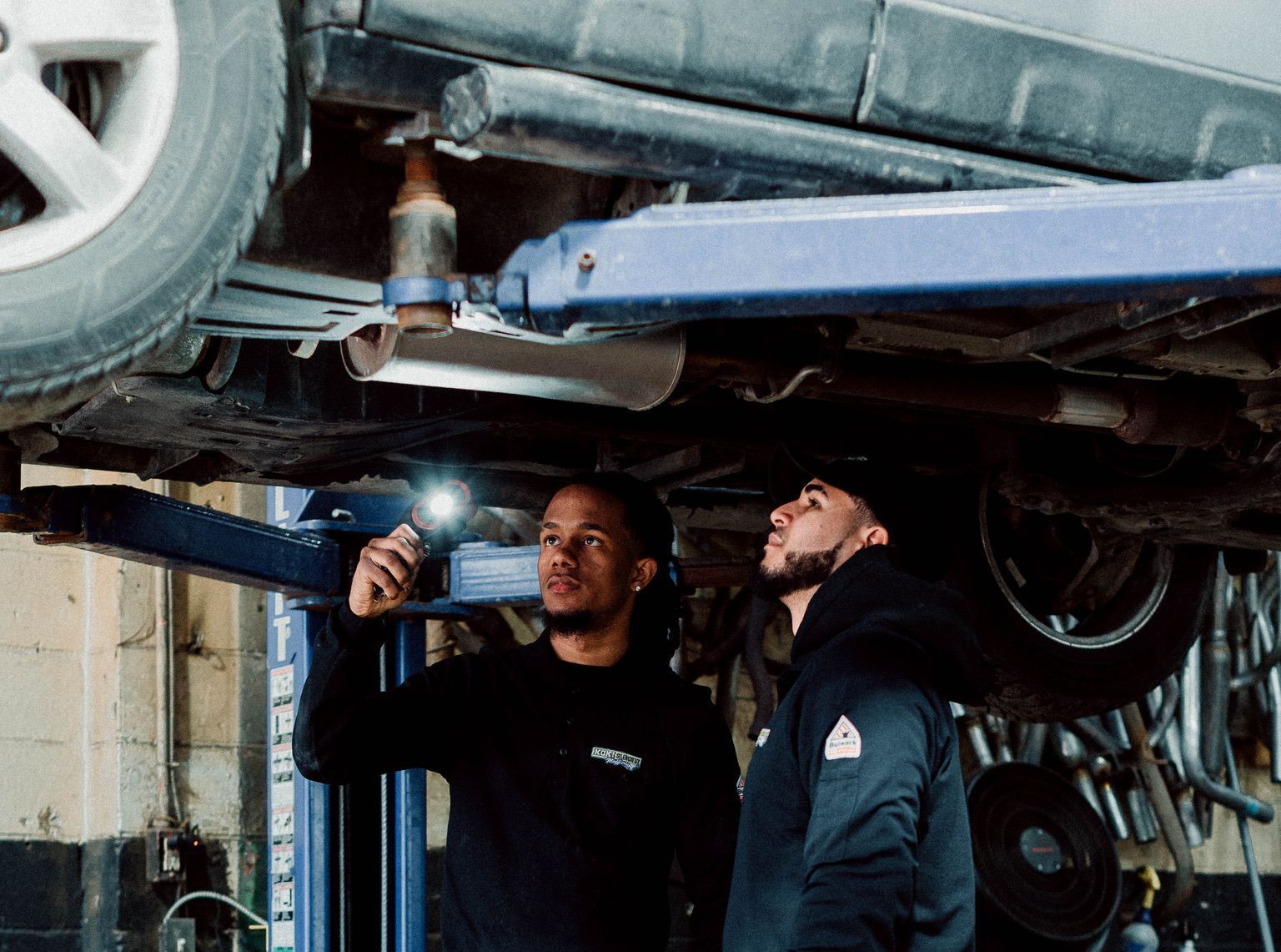Two men are working under a car on a lift in a garage.