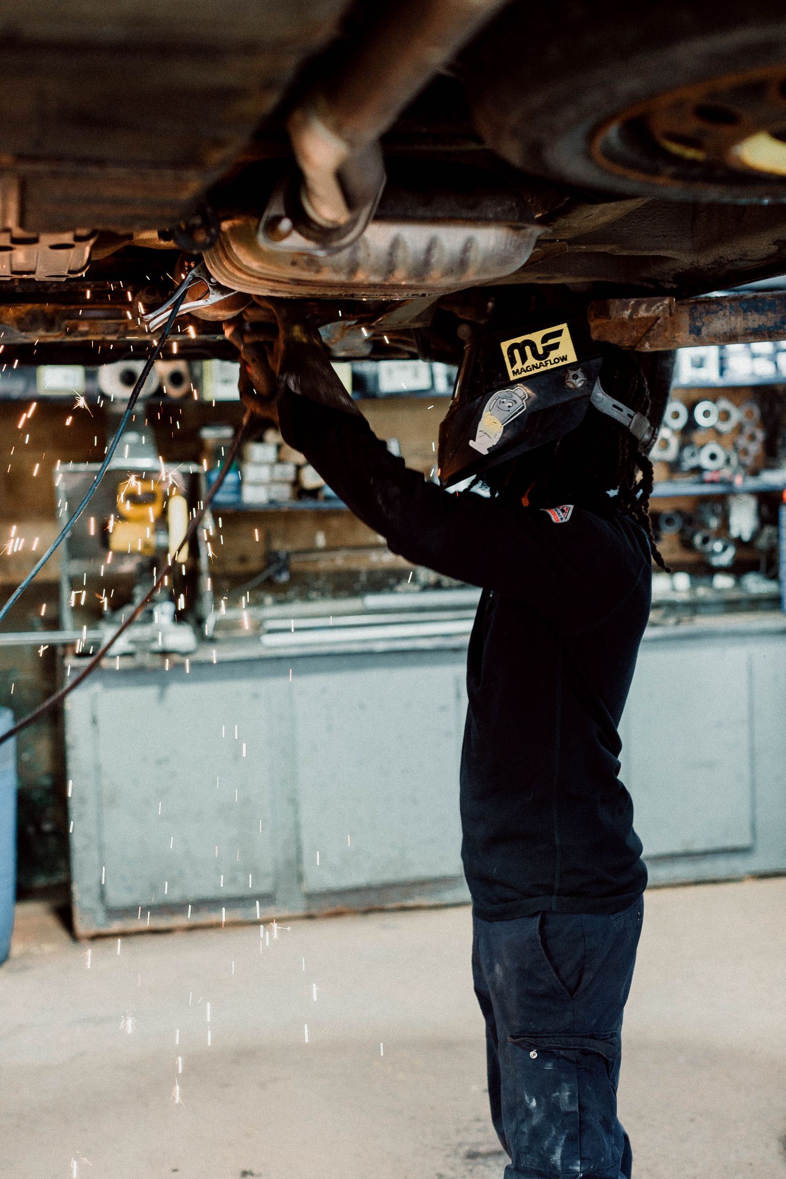 A man wearing a welding helmet is working under a car.