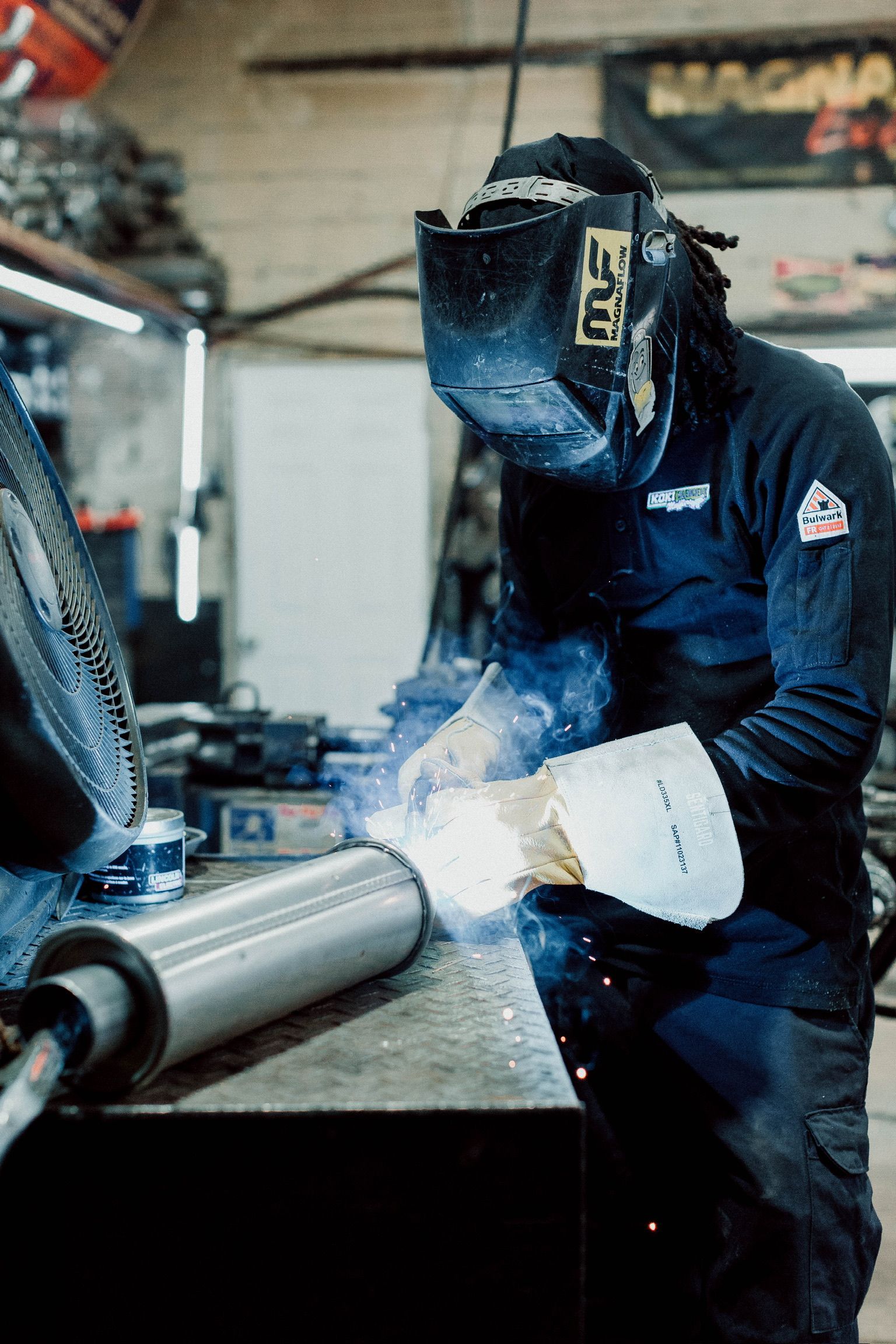 A man wearing a welding mask is welding a pipe in a garage.
