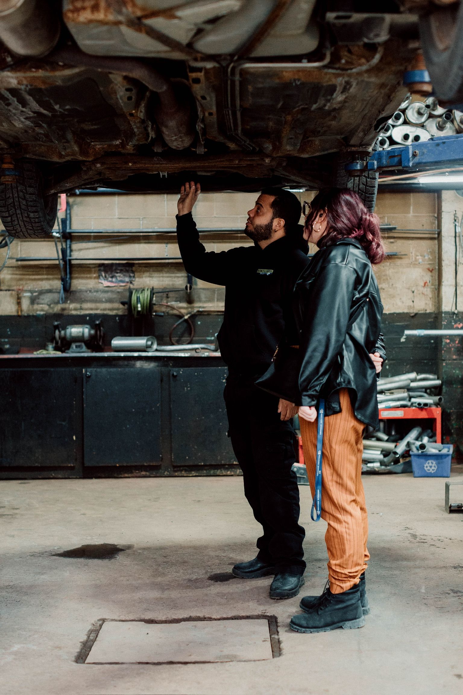 A man and a woman are looking under a car in a garage.