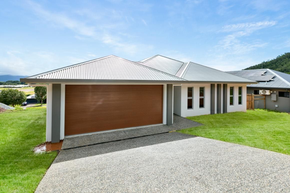A White House With a Brown Garage Door is Sitting on Top of a Lush Green Hillside — Ashlee Jones Homes in Gordonvale, QLD