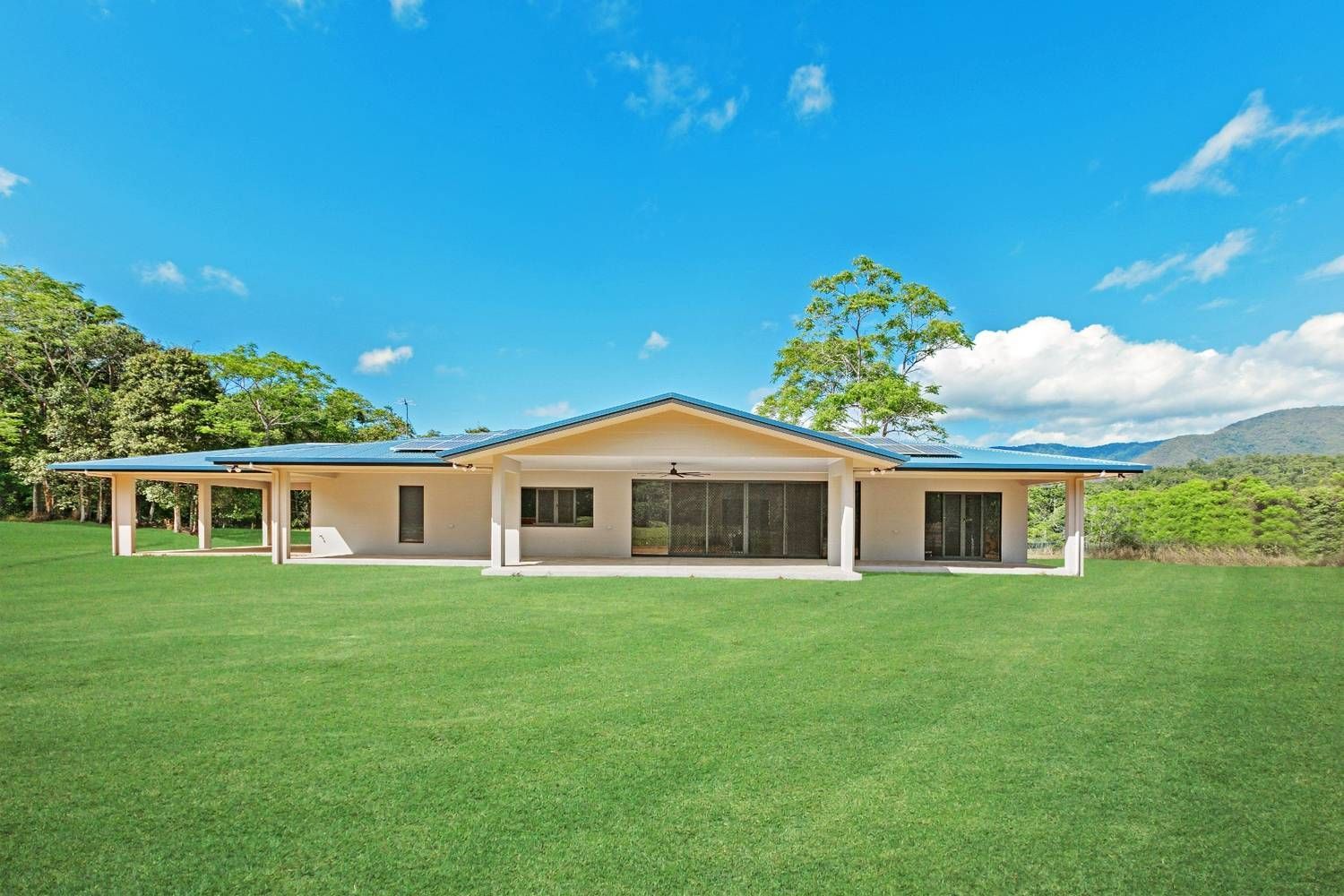 A White House With a Blue Roof is Sitting on Top of a Lush Green Field — Ashlee Jones Homes in Gordonvale, QLD