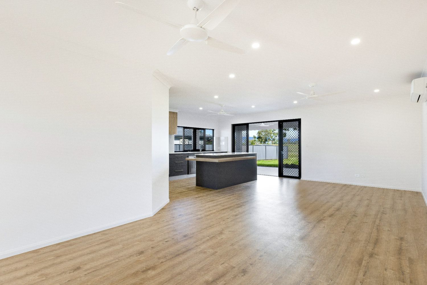 An Empty Living Room With Hardwood Floors and a Kitchen in the Background — Ashlee Jones Homes in Gordonvale, QLD
