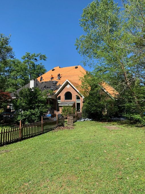 House with partially removed roof, construction in progress. Surrounded by green trees and grass, clear blue sky.