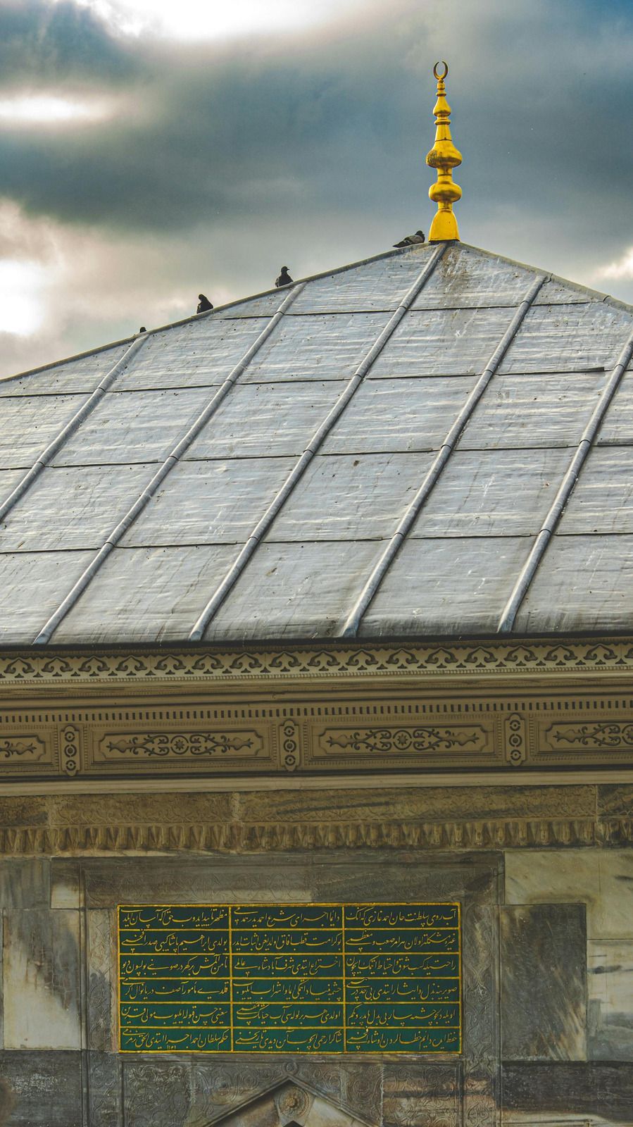 Stone building roof with golden finial under cloudy sky.