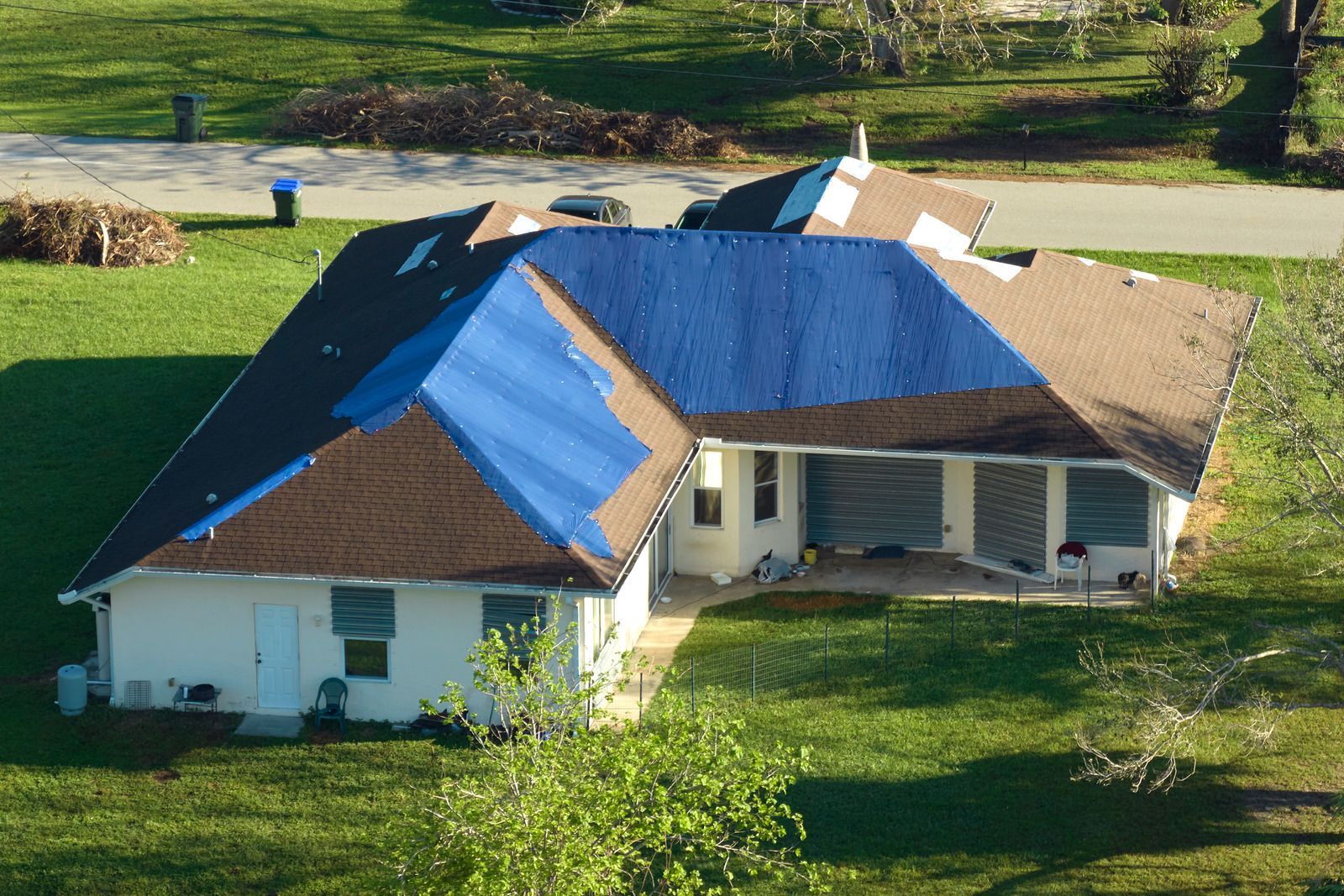 House with brown shingles and blue tarp covering damaged roof. Green lawn surrounds the home.