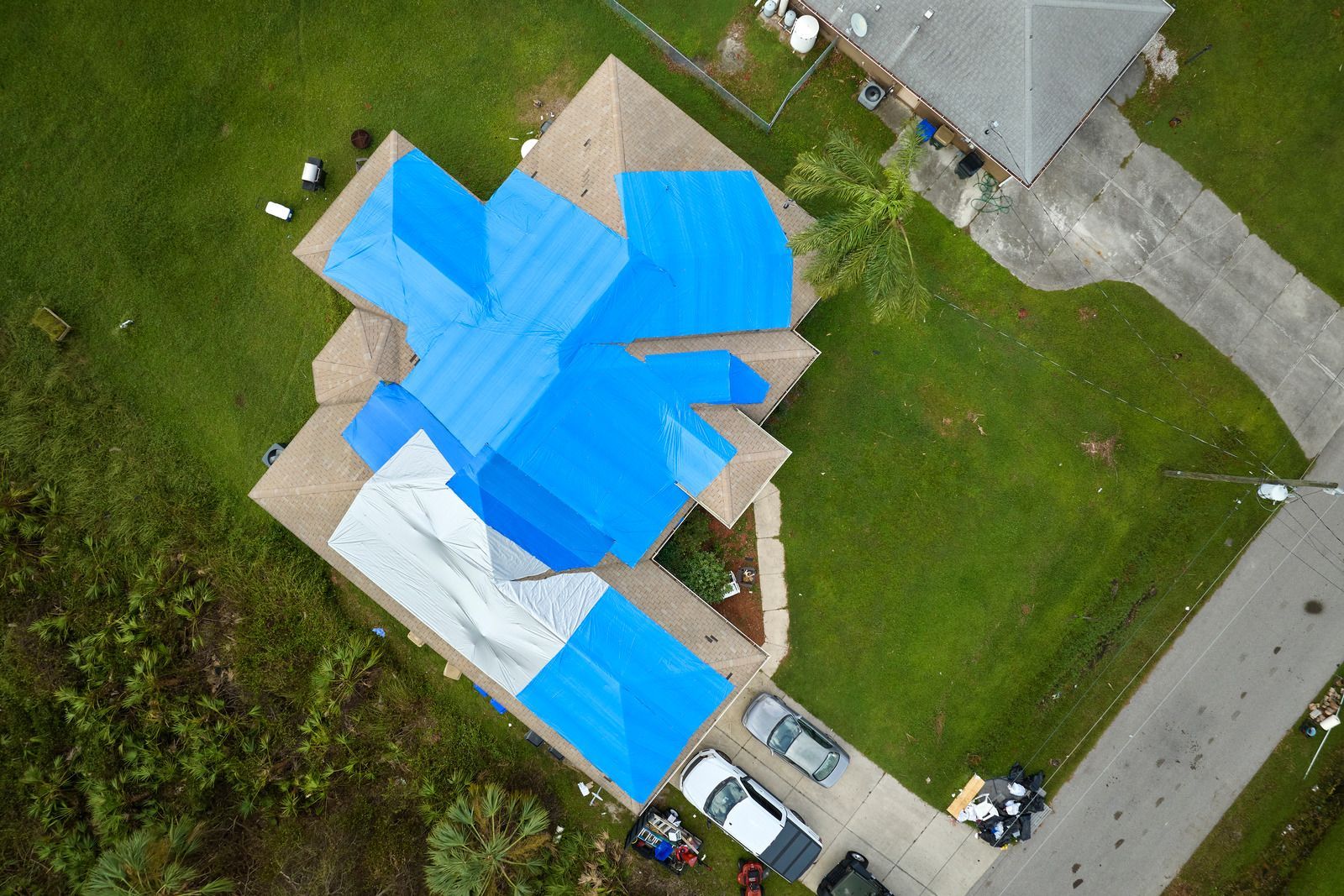 Aerial view of a house with sections of roof covered in blue and white tarps after a storm.