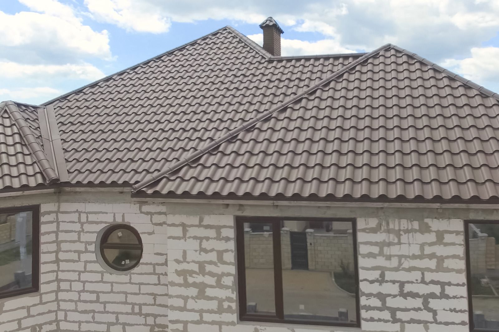 Brown metal tile roof of a house with chimneys, overlooking a field and another house.