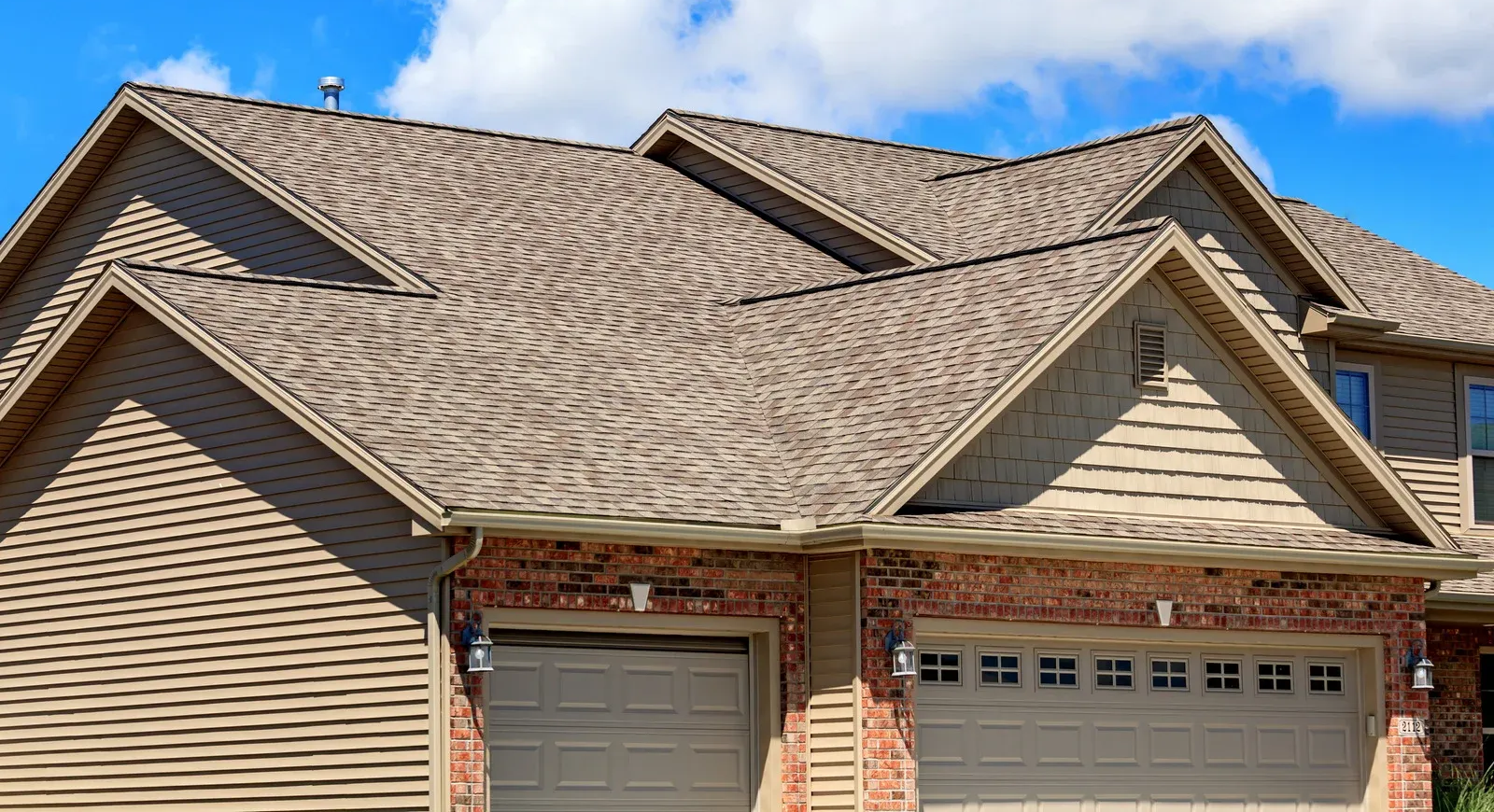 Tan house with brown roof and two garage doors, set against a blue sky with clouds.